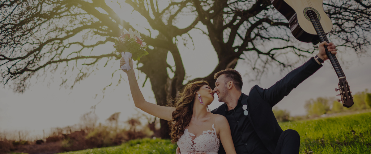 Symbolbild für Musik und Hochzeitsplanung: Paar in Hochzeitskleidung sitzt auf einer Wiese vor einem großen Baum, Frau hält Blumenstrauß hoch, Mann hebt Gitarre und sie berühren sich mit der Nase.