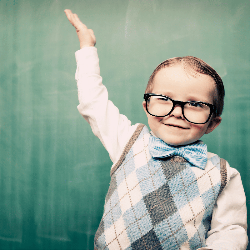 Kleiner Junge mit Brille, blauem Fliegen und argyle Strickweste, der vor einer grünen Tafel lachend eine Hand hebt