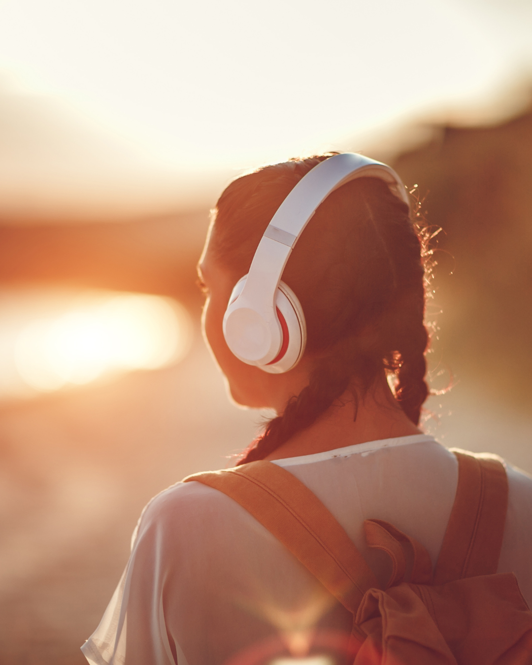 Rückansicht einer Person mit Zöpfen, die weiße Kopfhörer trägt und einen Rucksack auf den Schultern hat, vor einem Sonnenuntergang in warmer Lichtstimmung
