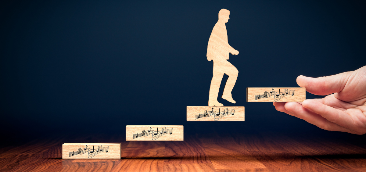 Motivation und Musik hängen eng zusammen: Holzfigur eines Mannes in Anzug, die auf Holzklötzen mit Musiknoten als Schritte nach oben geht, mit einer Hand, die den letzten Schritt hält, vor dunklem Hintergrund auf Holzboden.