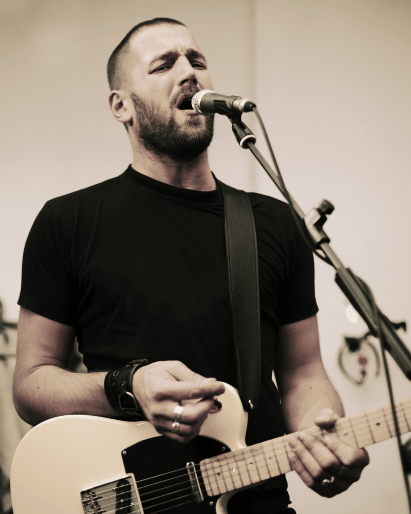Mann mit kurzem Haar und Bart, der mit geschlossenen Augen in ein Mikrofon singt und eine elektrische Gitarre spielt, trägt schwarzes T-Shirt und Lederarmband.