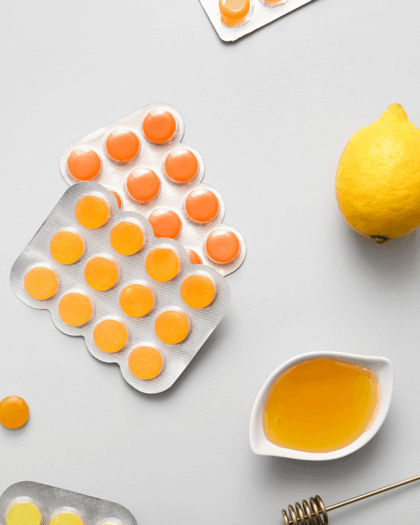 Stimmgesundheit und Medikamente: Blisterpackungen mit orangen und gelben Lutschtabletten auf grauem Hintergrund, daneben eine ganze Zitrone und ein kleines weißes Schälchen mit Honig sowie ein Honiglöffel.