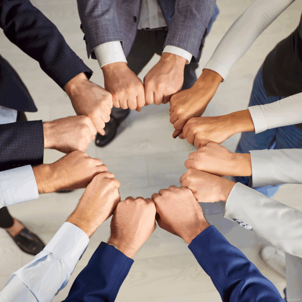 Team-Mitglieder bilden mit ihren Händen einen Kreis – Sinnbild für Zusammenhalt und Gemeinschaft durch Musik bei Firmenfeiern. Menschen verschiedener Hautfarben und mit Anzügen und Hemden stehen im Kreis und berühren mit ihren Fäusten in der Mitte die Fäuste der anderen als Zeichen von Teamgeist und Zusammenarbeit.