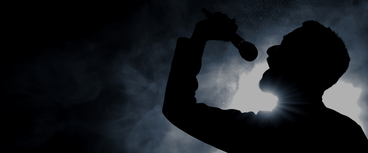 Symbolbild für Stimmgesundheit: Schattenriss eines Mannes, der mit erhobenem Arm in ein Mikrofon singt, vor hellem Licht und Nebel im Hintergrund.