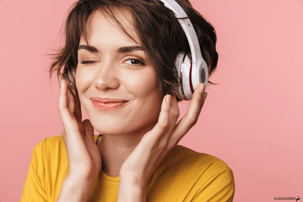 Symbolbild Musik und Emotionen: Junge Frau mit kurzen braunen Haaren trägt weiße Kopfhörer, trägt ein gelbes Shirt, hält die Kopfhörer mit beiden Händen und zwinkert mit lachendem Gesicht vor einem rosa Hintergrund.