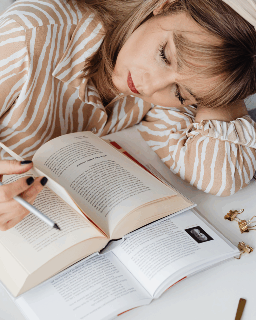 Lernen mit Musik kann auch Ablenkung sein. Daher sollte man auf die richtige Musikauswahl achten.Frau mit gestreiftem Hemd, die mit geschlossenen Augen auf einem Schreibtisch liegt und ein Buch mit einem Stift in der Hand liest, umgeben von geöffneten Büchern und Büroklammern.