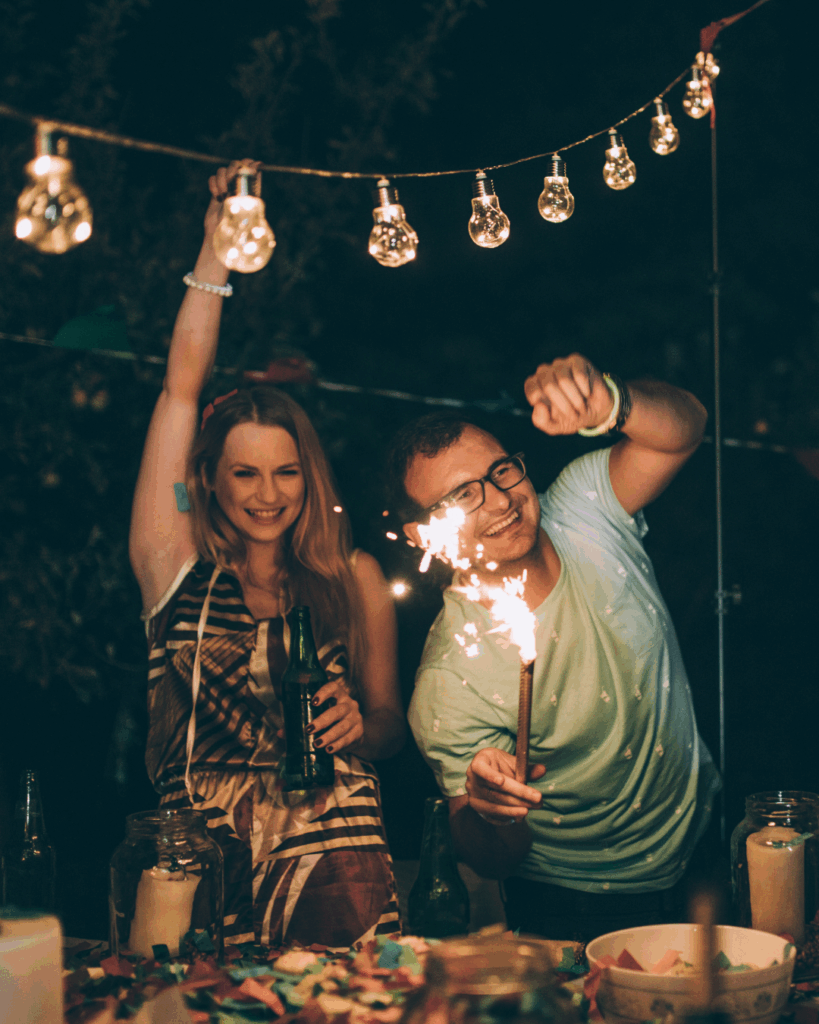 Zwei Menschen feiern abends unter Girlande mit Glühbirnen, eine Frau hält eine Flasche und hebt den Arm, ein Mann trägt eine Brille und hält eine Wunderkerze, auf dem Tisch sind bunte Konfetti und dekorierte Kerzen in Gläsern.