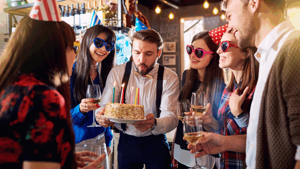 Ob Live-Musik oder DJ - finde die perfekte Musik für deine Geburtstagsfeier und lass die Party beginnen! Eine Gruppe von jungen Erwachsenen mit Partyhüten und Herzförmigen Sonnenbrillen feiert in einem gemütlichen Raum, während ein Mann in der Mitte Kerzen auf einem Geburtstagskuchen auspustet und die anderen mit Weingläsern anstoßen und lächeln.