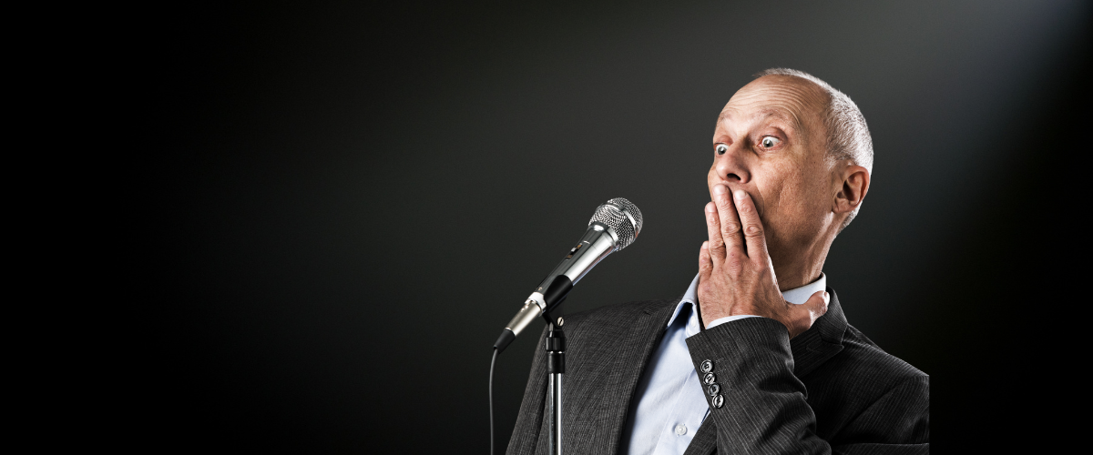 Umgang mit Lampenfieber: Mann mit Anzug und hellblauem Hemd steht vor einem Mikrofon und hält überrascht die Hand vor den Mund vor dunklem Hintergrund