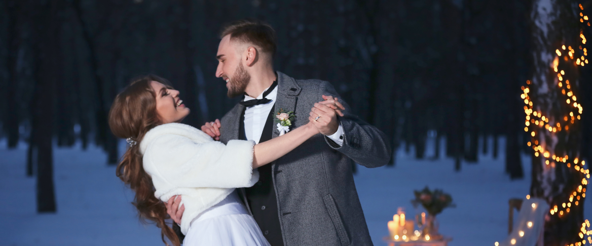Winterhochzeit: Braut und Bräutigam tanzen lachend im Schnee in einem Wald bei Nacht, umgeben von Kerzenlicht und Lichterketten an einem Baum.