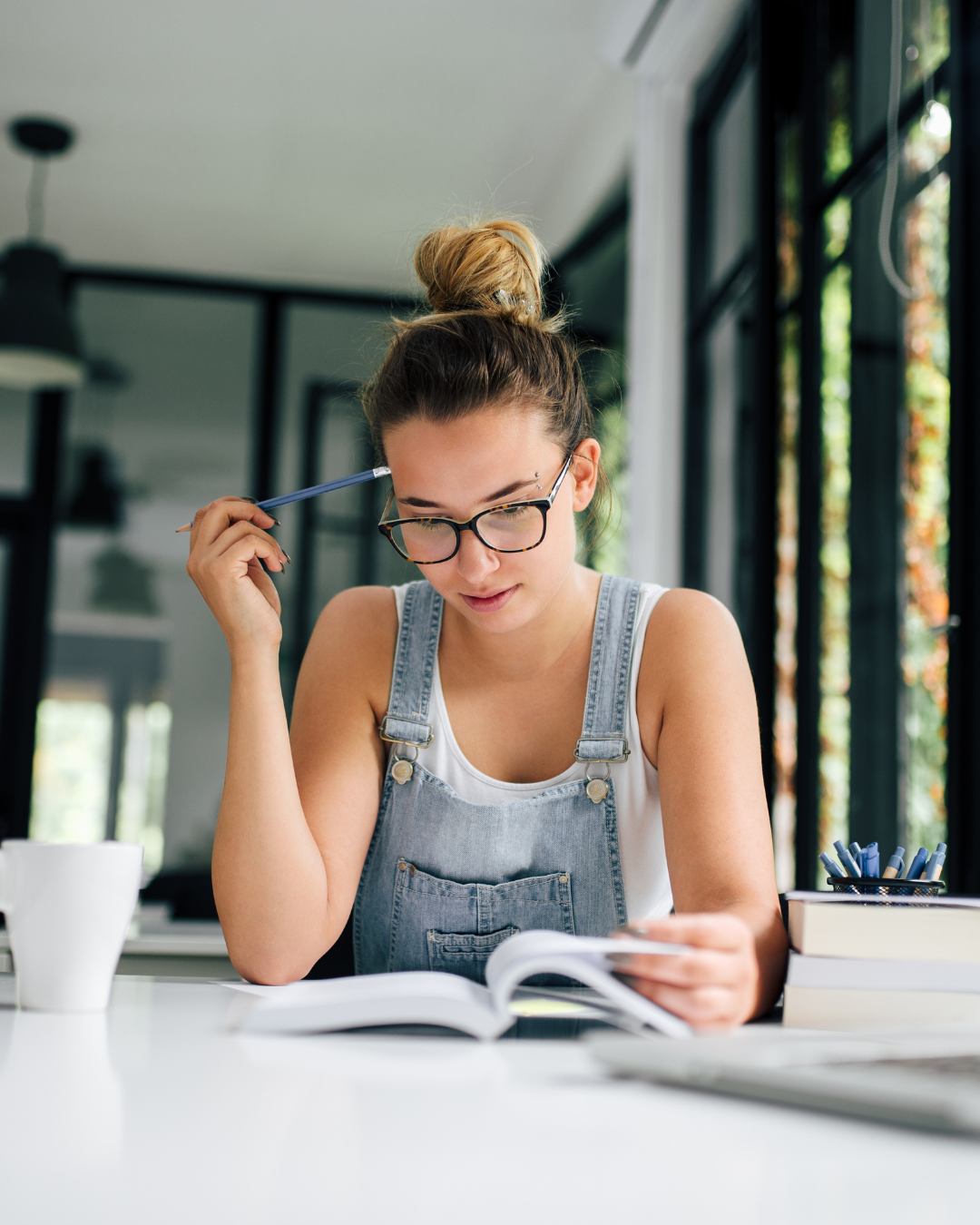 Junge Frau mit Brille und hochgebundenem Haar liest konzentriert ein Buch an einem weißen Tisch in einem hellen Raum, sie hält einen Stift an ihre Stirn und trägt eine Jeans-Latzhose über einem weißen Tanktop.
