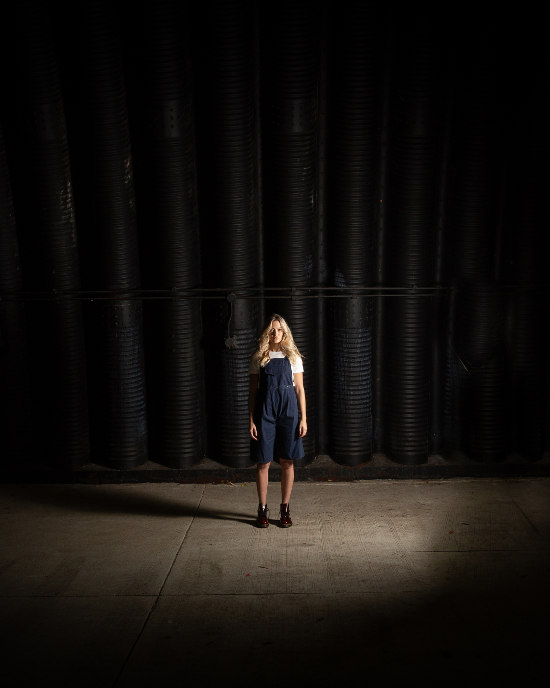 Lampenfieber muss keine Schwäche sein! Symbolbild: Eine Frau mit blonden Haaren, die ein weißes T-Shirt, eine dunkelblaue Latzhose und dunkle Stiefel trägt, steht allein in einem spärlich beleuchteten, dunklen Raum vor einer Wand mit großen schwarzen Rohren.