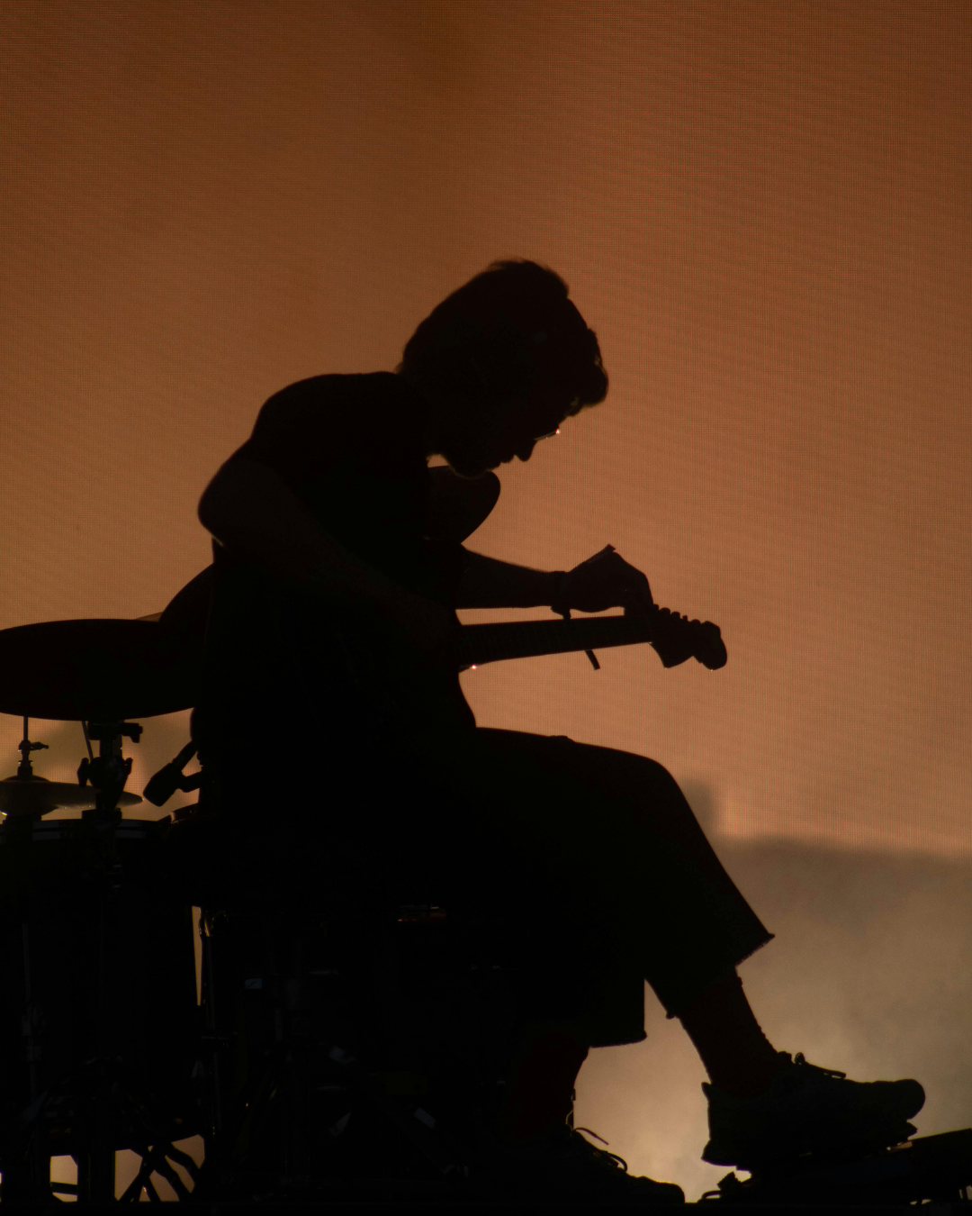 Lampenfieber und Nervosität: Silhouette einer sitzenden Person, die eine Gitarre stimmt, vor einem orangefarbenen Hintergrund mit unscharfen Trommeln im Hintergrund.