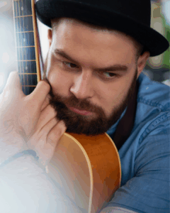 Nahaufnahme eines bärtigen Mannes mit schwarzem Hut, der nachdenklich eine akustische Gitarre umarmt und in die Ferne schaut, er trägt ein blaues Hemd.