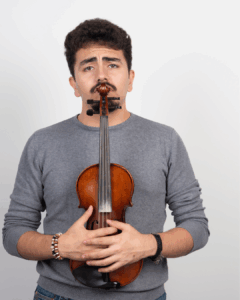 Mann mit dunklem lockigem Haar und Schnurrbart, der eine Violine vor sich hält, die an sein Gesicht gehalten wird, er trägt einen grauen Pullover und Armbänder, vor neutralem Hintergrund.