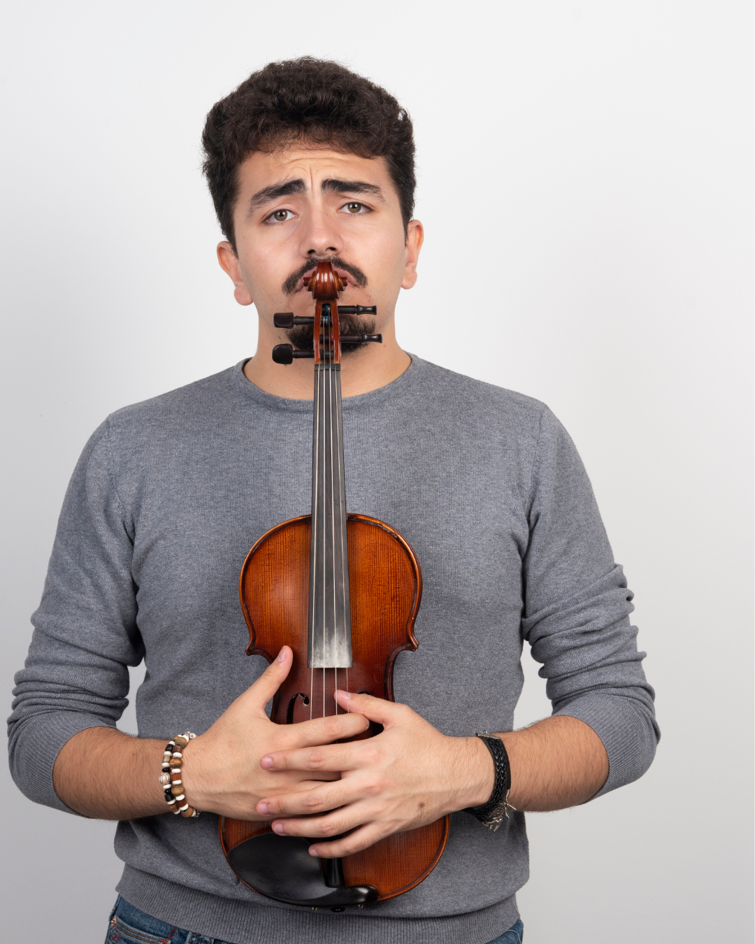 Mann mit dunklem lockigem Haar und Schnurrbart, der eine Violine vor sich hält, die an sein Gesicht gehalten wird, er trägt einen grauen Pullover und Armbänder, vor neutralem Hintergrund.