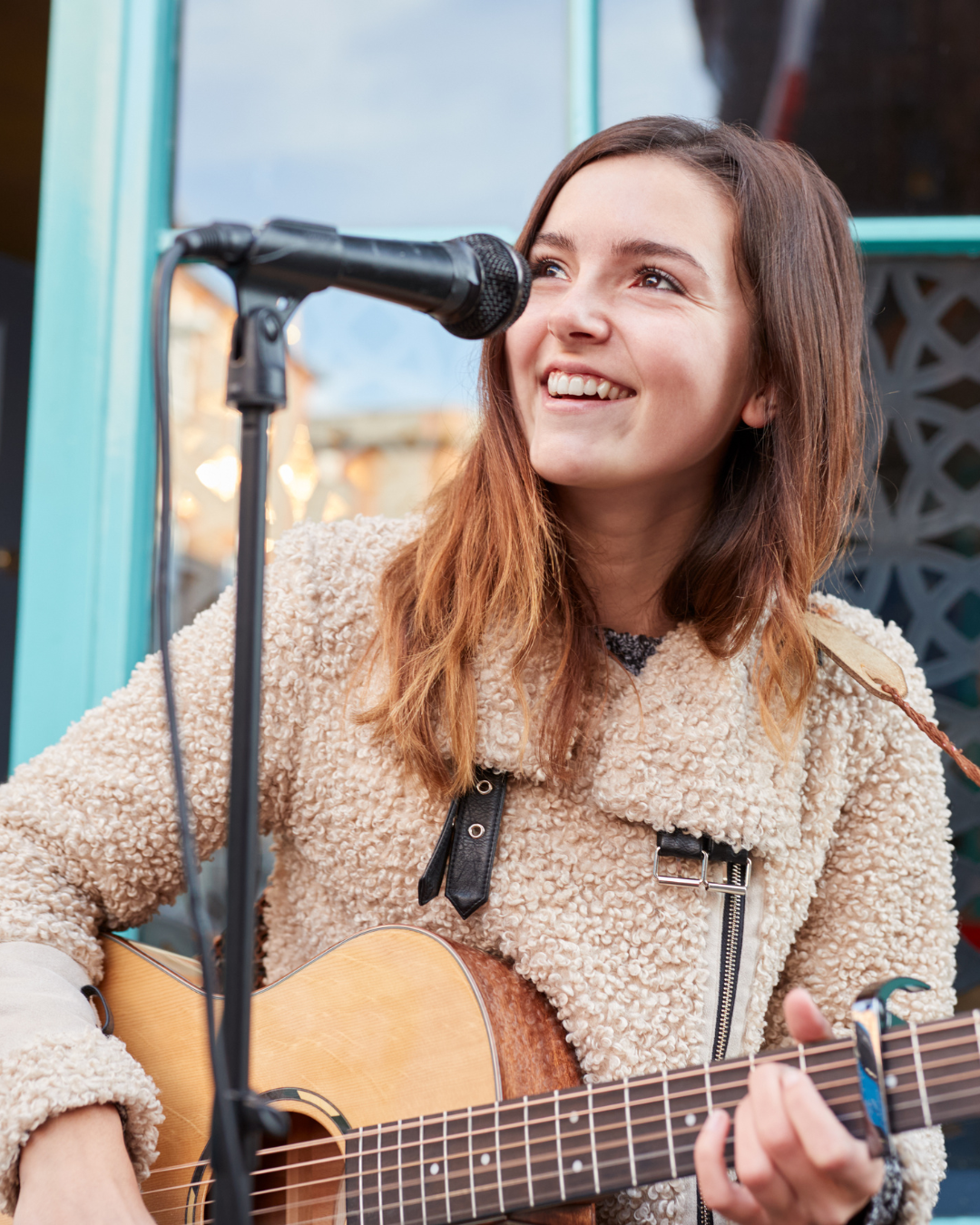Lampenfieber für sich nutzen: Junge Frau mit hellbraunem lockigem Mantel, die lächelnd eine Akustikgitarre spielt und vor einem Mikrofon in einer urbanen Umgebung steht.