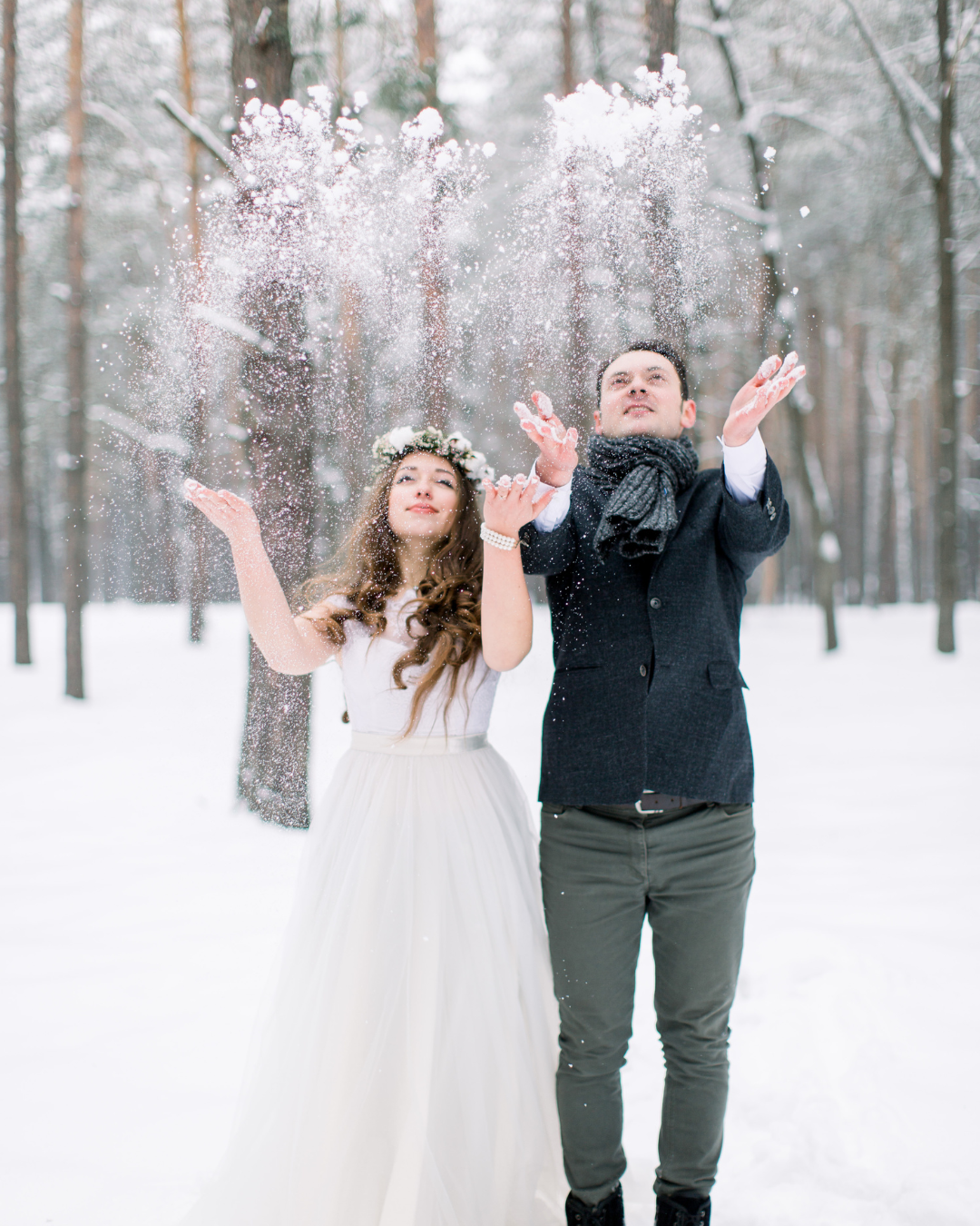Die perfekte Winterhochzeit: Paar im Winterwald, Frau in weißem Kleid mit Blumenkranz und Mann in dunklem Mantel werfen Schnee in die Luft, umgeben von schneebedeckten Bäumen.