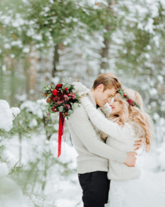 Braut und Bräutigam umarmen sich im verschneiten Wald, mit winterlichem Blumenstrauß in Bordeaux-Tönen.