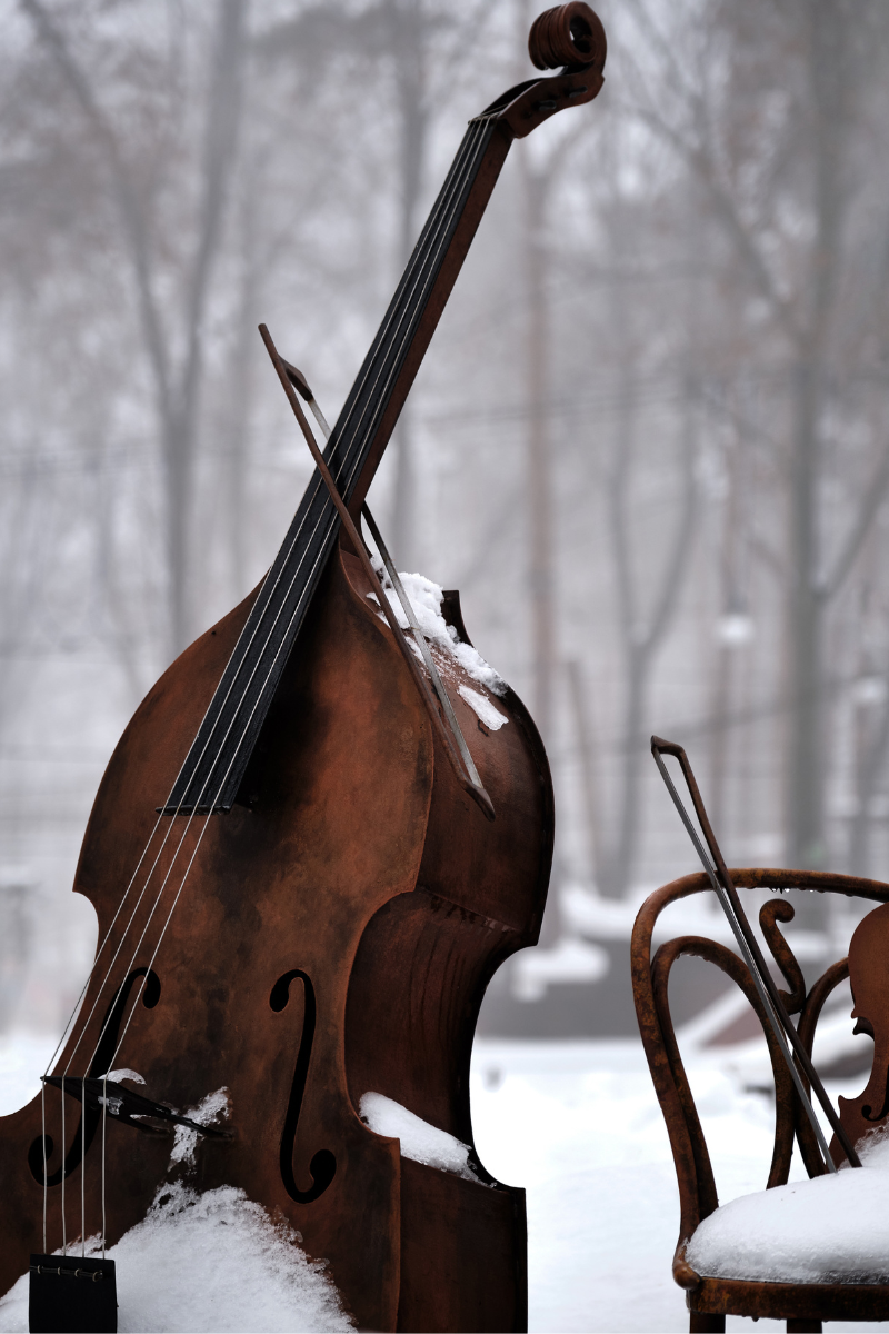 Kontrabass mit Schnee bedeckt, stehend im Freien vor einem verschwommenen winterlichen Hintergrund mit Bäumen, daneben ein alter Stuhl mit Schneeschicht und einem weiteren Streichinstrument darauf.