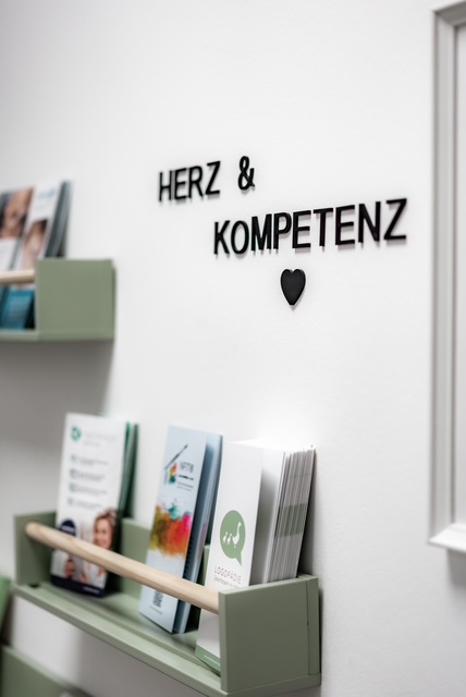 Das Schnatterwerk - Praxis für Logopädie: Wand mit schwarzem Schriftzug "HERZ & KOMPETENZ" und Herzsymbol darüber, darunter grüne Wandregale mit verschiedenen Broschüren und Informationsmaterialien.