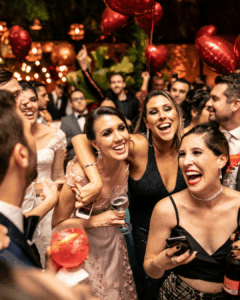 Fröhliche Menschen in festlicher Kleidung bei einer Party oder Feier mit roten Luftballons und Getränken, umgeben von warmem Lichterglanz und einer lebhaften Atmosphäre.