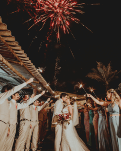 Hochzeitsmusik zum Hochzeitstanz: Hochzeitspaar küsst sich draußen bei Nacht, umgeben von Gästen, die Wunderkerzen halten, während über ihnen rote Feuerwerke am Himmel explodieren.