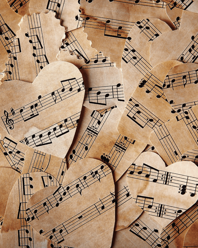 Musik als Herz unserer Gesellschaft. Mehrere herzförmige Papierschnipsel mit gedruckten Notenlinien und Musiknoten, übereinander liegend auf braunem Hintergrund.