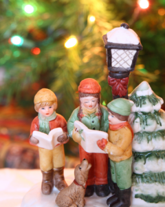 Singende Kinderfiguren zur Weihnachtszeit – Gemeinschaft und Musik erleben - Figuren von drei Kindern mit Weihnachtsmützen und Büchern, sitzender Hund, Laterne und verschneiter Tannenbaum im Hintergrund vor Weihnachtsbaumlicht.