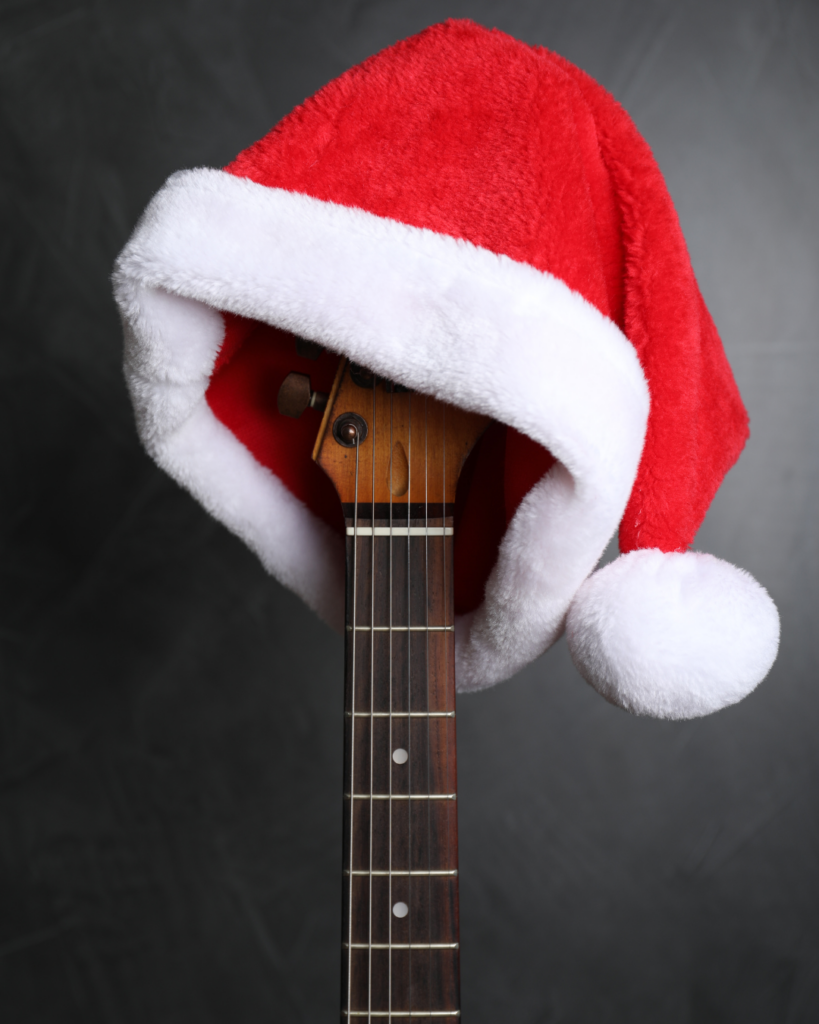 Gitarre mit Weihnachtsmütze – moderne Weihnachtsmusik im Zeitalter der Medien - Gitarrenhals mit weihnachtlicher roter Santa-Mütze mit weißem Plüschrand und Bommel auf dunklem Hintergrund.