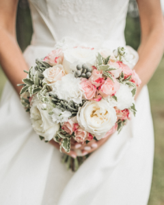 Braut hält einen Brautstrauß mit weißen und rosa Rosen und grünen Blättern vor weißem Hochzeitskleid.