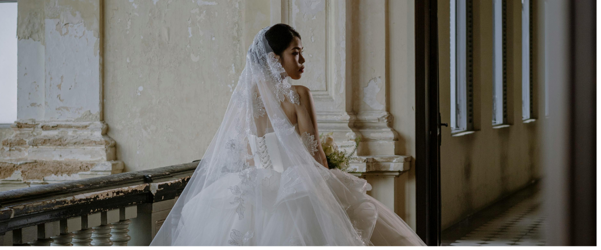 Einzug der Braut: Braut im weißen Hochzeitskleid mit langem Spitzen-Schleier sitzt an balustradengeländer in altem Gebäude mit marmoriertem Putz.
