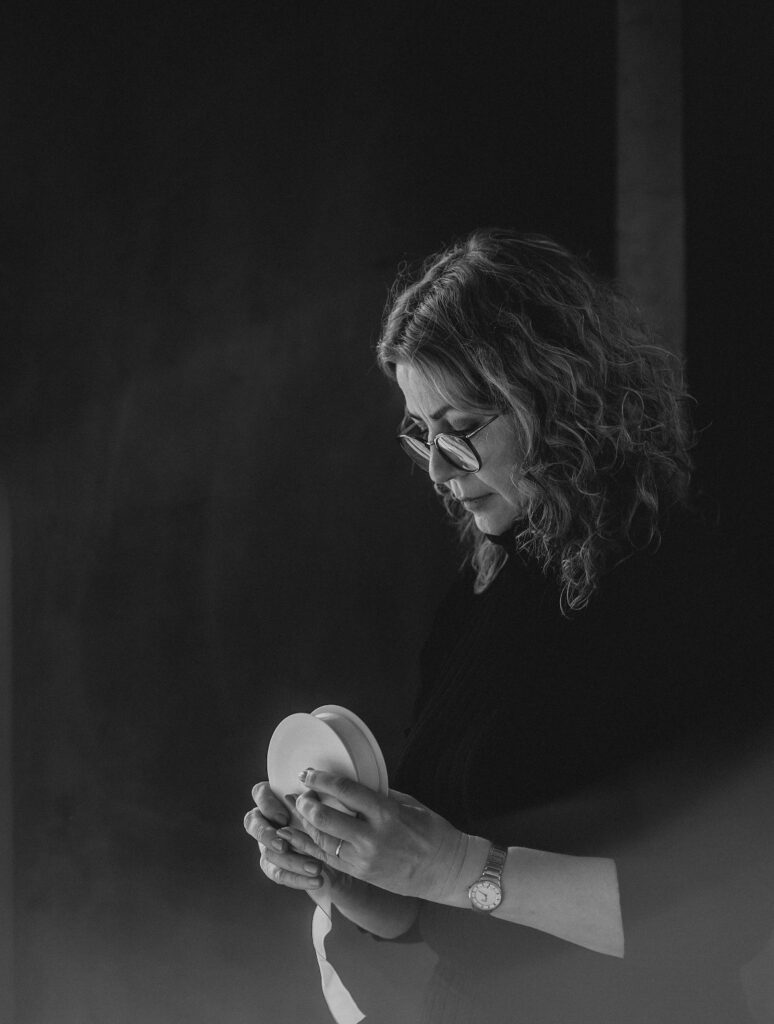 Schwarz-Weiß-Porträt einer Frau mit lockigem Haar und Brille, die ein Band von einer Spule abrollt und nach unten blickt, vor dunklem Hintergrund.