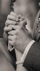 Schwarz-weiß-Foto eines Paares, das sich an den Händen hält, beide tragen Eheringe, die Frau trägt ein Perlenarmband, Nahaufnahme mit Fokus auf den Händen und einem lächelnden Gesicht im Hintergrund.