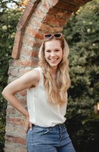 Junge blonde Frau mit langen Haaren, weißer Bluse und blauer Jeans steht lächelnd vor einem gemauerten Steinbogen im Freien mit grüner Bepflanzung im Hintergrund.