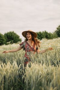 Frau mit Strohhut und gemustertem Kleid, die lächelnd mit ausgestreckten Armen in einem Weizenfeld steht, umgeben von Blüten und Bäumen im Hintergrund.