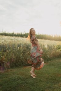 Junge Frau in einem gemusterten, fließenden Kleid tanzt barfuß auf einer Wiese bei Sonnenuntergang mit strahlendem Gesichtsausdruck.