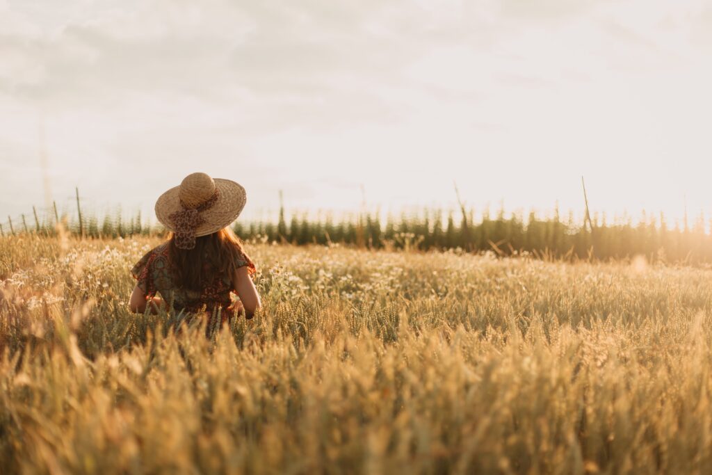 Frau mit Strohhut und langen Haaren steht in einem goldenen Getreidefeld bei Sonnenuntergang, Blick in die Ferne.