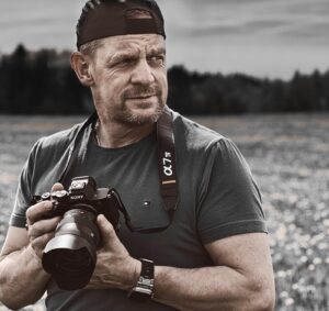 Mittelalter Mann mit schwarzer Baseballkappe rückwärts, dunkelblauem T-Shirt und Armbanduhr hält eine Sony α7 IV Kamera und schaut nach rechts, im Hintergrund unscharfes Feld mit Bäumen