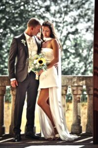 Braut und Bräutigam in Hochzeitskleidung stehen eng umschlungen auf einem Balkon mit Steingeländer, die Braut hält einen Strauß mit Sonnenblume.