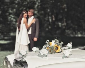Brautpaar umarmt sich verschwommen im Hintergrund, im Vordergrund ein weißes Hochzeitsauto mit weißen Rosen und einem Strauß mit Sonnenblumen dekoriert, an einem sonnigen Tag im Freien