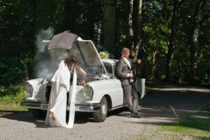 Braut in weißem Kleid und Bräutigam im Anzug neben einem alten weißen Auto mit geöffneter Motorhaube, die Braut versucht den Motor zu reparieren, während Rauch aus dem Motorraum aufsteigt, der Bräutigam schaut auf die Uhr und hält eine Flasche, Umgebung ist ein bewaldeter Weg mit Sonnenlicht und Schatten