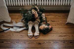 Braune Damen-Sandaletten, Rosenstrauß und Schmuckschatulle mit Blumen auf Holzboden vor Heizkörper in Raum mit weißen Vorhängen.