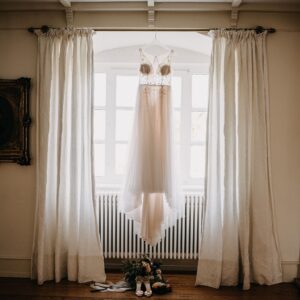 Elegantes Hochzeitskleid mit Spitze und Tüll, an einem Fenster zwischen weißen Vorhängen aufgehängt, darunter Brautschuhe und Blumenstrauß.