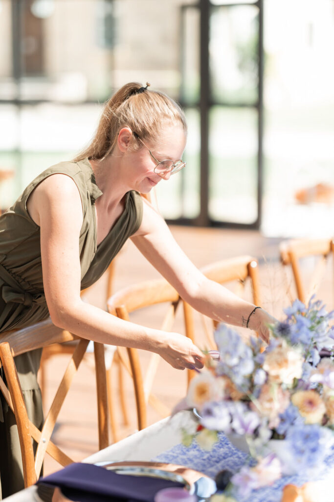 Frau mit Brille und Zopf richtet mit einem Lächeln den Tisch mit Blumendekoration in hellem Raum ein.