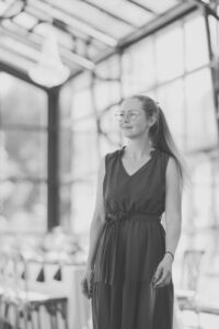 Frau mit Brille und langem Pferdeschwanz in ärmellosem Kleid steht in hellem Raum mit hohen Fenstern.