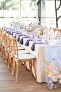 Festlich gedeckter langer Tisch mit weißen Hussen, lila Servietten, blumiger Dekoration und Holzboden in hellem Raum