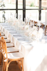 Langer festlich gedeckter Tisch mit weißen Blumen, Kerzen und Holzstühlen in hell erleuchtetem Raum.