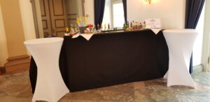 Eine Cocktailbar mit schwarzem Tischtuch, auf dem verschiedene Flaschen und bunte Strohhalme stehen, flankiert von zwei hohen weißen Stehtischen in einem Raum mit Marmorboden und dunklen Vorhängen.