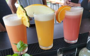 Drei bunte Cocktailgläser mit Schaum, von links nach rechts dekoriert mit Ananas, Honigmelone und Orangenscheibe, stehen nebeneinander auf einem schwarzen Barpad.